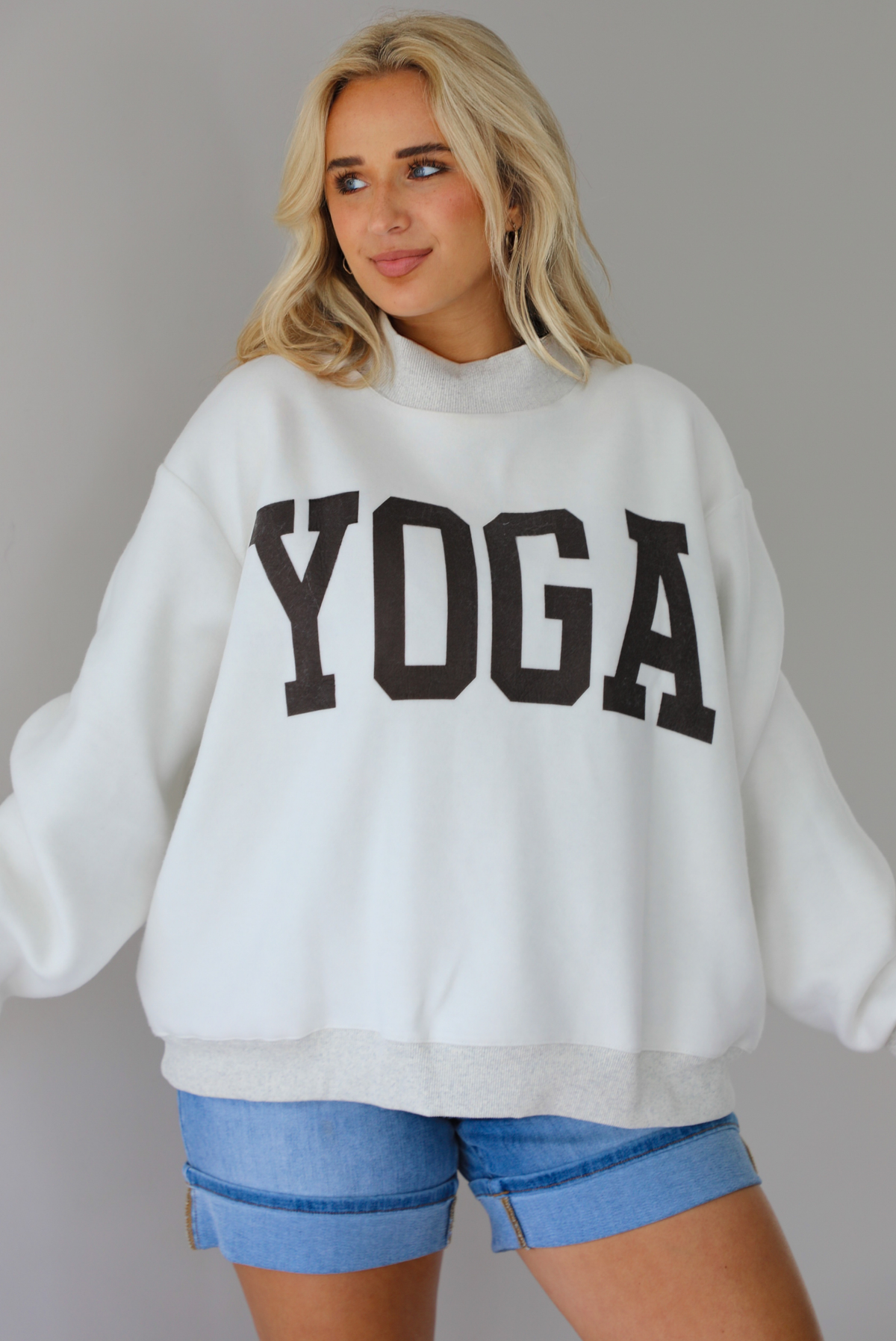 Woman wearing a heather grey oversized sweatshirt, reversed inside out, with the word 'yoga' written in collegiate font on the front with medium wash denim shorts indoors against a white wall.