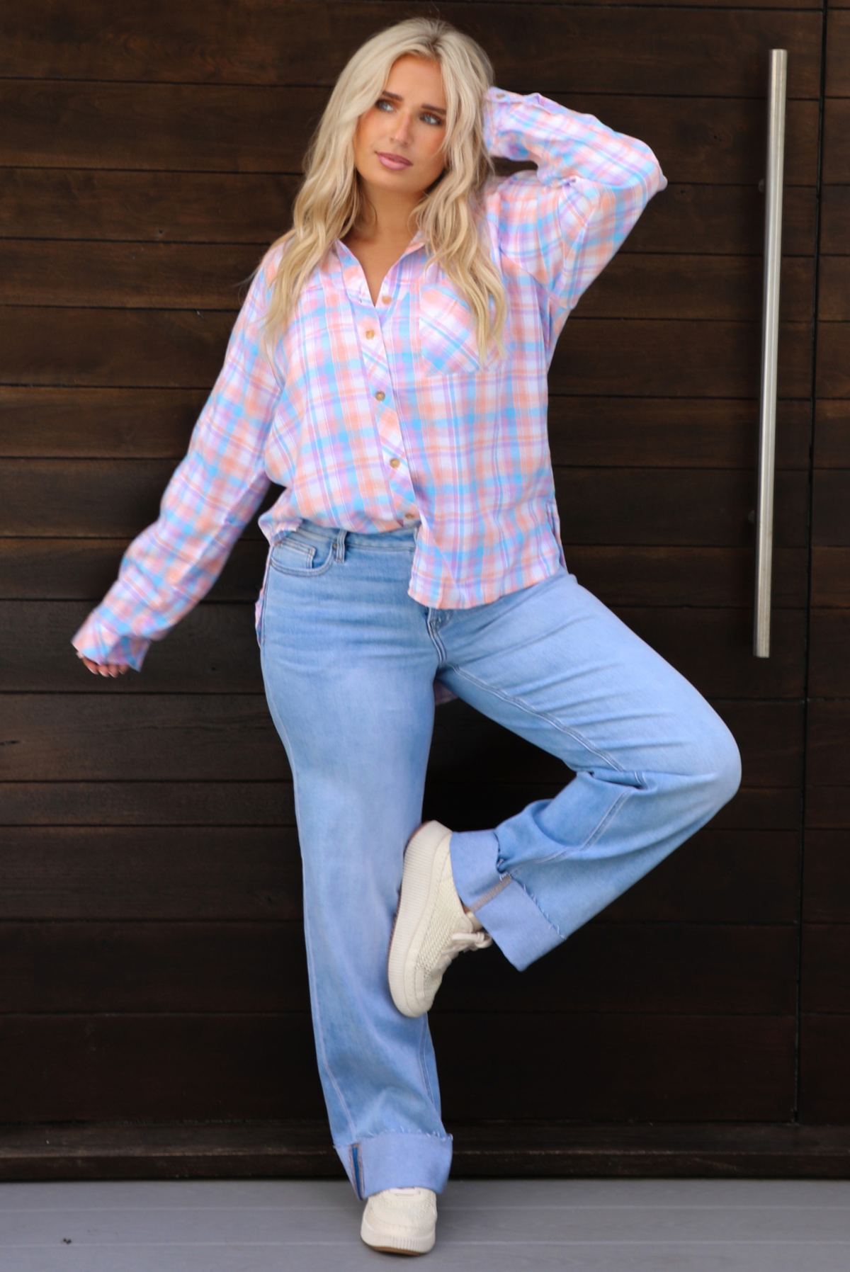 A woman wearing a pink, blue, and purple plaid print pattern throughout that has a collared neckline, functional button closures along the front, and an oversized fit, with one side tucked into light wash denim that have cuffed hems, standing in front of a dark brown wooden door. 