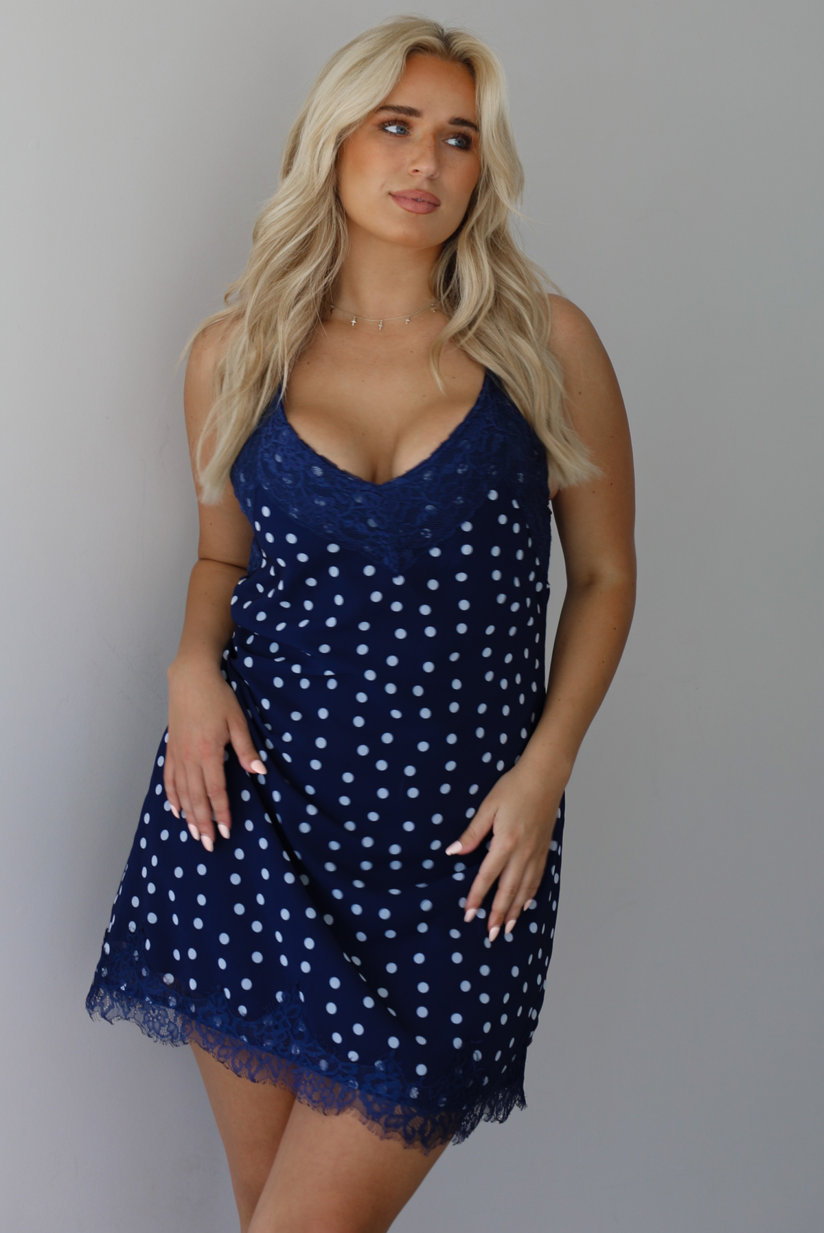 Woman wearing a navy dress with a white polka dot print pattern throughout with blue lace detailing at the neckline and hem indoors against a white wall.