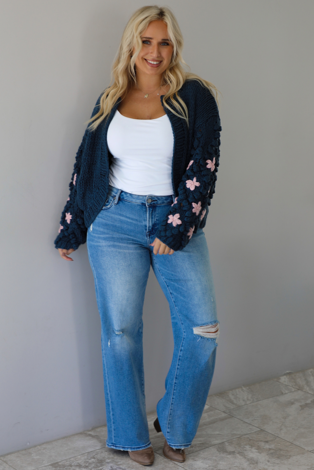 A woman wearing a navy oversized cardigan with a pink embroidered floral design on the sleeves, paired with a white tank, mid-rise medium wash denim jeans, standing indoors against a white wall with white marble flooring. 