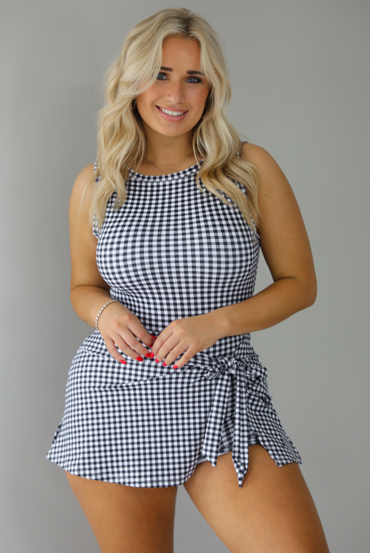 Woman wearing a black and white gingham print athletic romper that has a wrap-style skirt detail. The photo is taken indoors against a white wall.