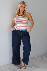 A woman modeling a sleeveless ribbed knit top with horizontal stripes in red, light blue, navy, and white, paired with dark wide-leg jeans and metallic rose gold flats, standing indoors against a white wall with white marble flooring. 