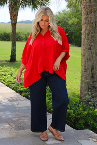 A woman wearing a bright red oversized short-sleeve blouse and dark wide-leg jeans, standing outdoors on a stone walkway with trees and green grass in the background.