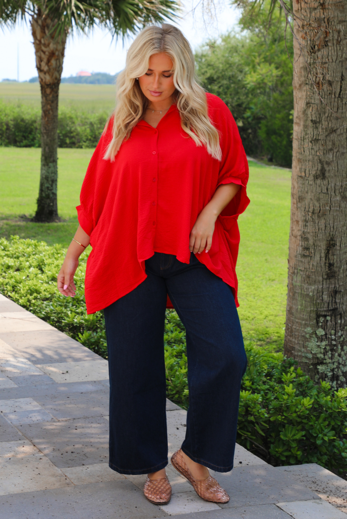 A woman wearing a bright red oversized short-sleeve blouse and dark wide-leg jeans, standing outdoors on a stone walkway with trees and green grass in the background.