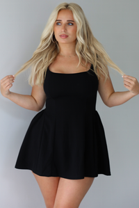 Woman wearing a black mini dress with thick straps, a fitted bodice, and flowy skirt indoors against a white wall.
