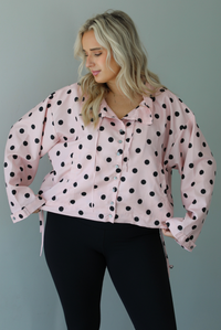 A woman wearing a pink button-down jacket with a black polka dot print pattern, paired with black leggings, white ankle socks, and black slide on slippers, standing indoors against a white wall.