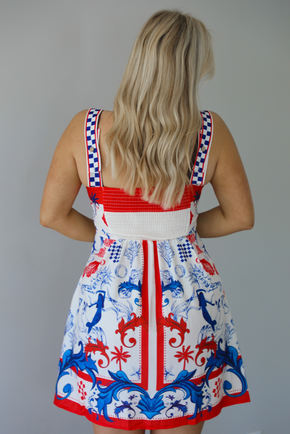 Woman wearing a white dress with red and blue sea themed graphics throughout with thick straps and functional pockets on either side. The photo is taken indoors against a white wall and shows the back of the dress with its smocked detailing. 