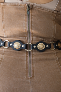 A closeup of the back of a woman wearing a taupe brown fitted midi dress, focusing in on the zipper closure in the back, and it also shows a closeup of the black belt that's worn at the waist. 