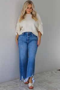 Woman wearing a cream colored quarter sleeve sweater that has ribbed details at the neckline, on the sleeves, and on the hem, with non-functional button closures on the sleeves. The sweater has a relaxed fit and is paired with a medium wash, high waisted, distressed hem denim with sand colored sandals. The photo is taken indoors against a white wall with white marble flooring. 