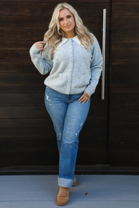 Woman model wearing a heather grey cardigan with a white lace bib at the neckline, has functional button closures in the front, functional pockets in the front, and a relaxed fit. The cardigan is paired with a medium wash denim and tan ugg boots. The photo is taken outdoors against a dark brown wooden door.