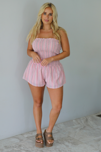 A woman wearing a pink and multi-colored vertically striped strapless romper, standing indoors against a white wall with white marble flooring. 