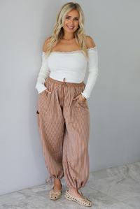A woman wearing a cream off the shoulder long sleeved top tucked into a pair of camel and cream striped pants, standing indoors against a white wall with white marble flooring. 