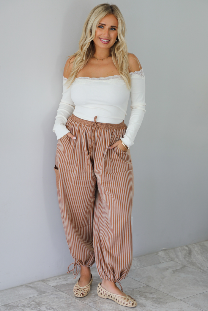 A woman wearing a cream off the shoulder long sleeved top tucked into a pair of camel and cream striped pants, standing indoors against a white wall with white marble flooring. 