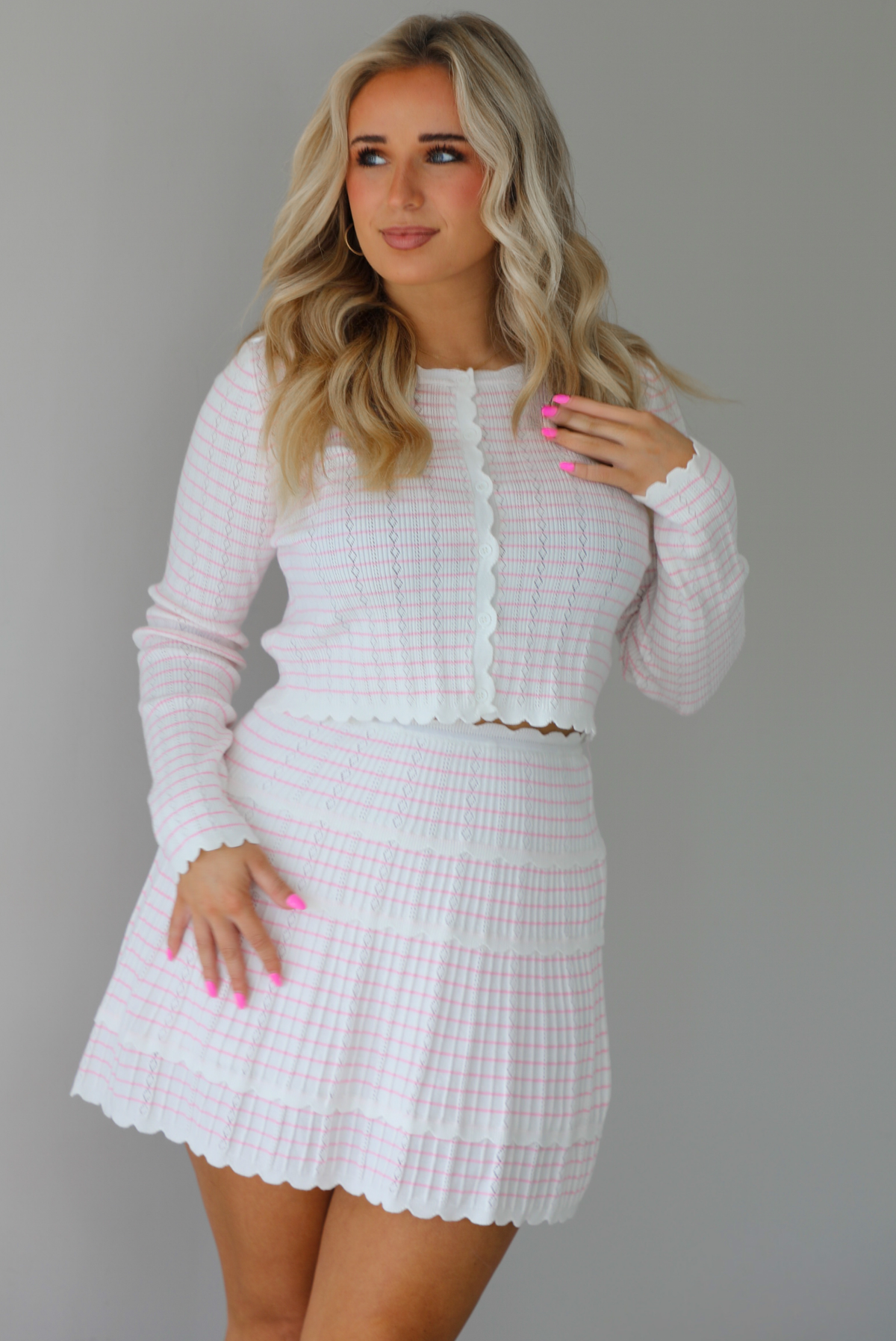 Woman wearing a two piece set that featuring a cropped cardigan and a high waisted skirt. The set is white with a pink horizontal striped pattern throughout, with scalloped hem details. The photo is taken indoors against a white wall.