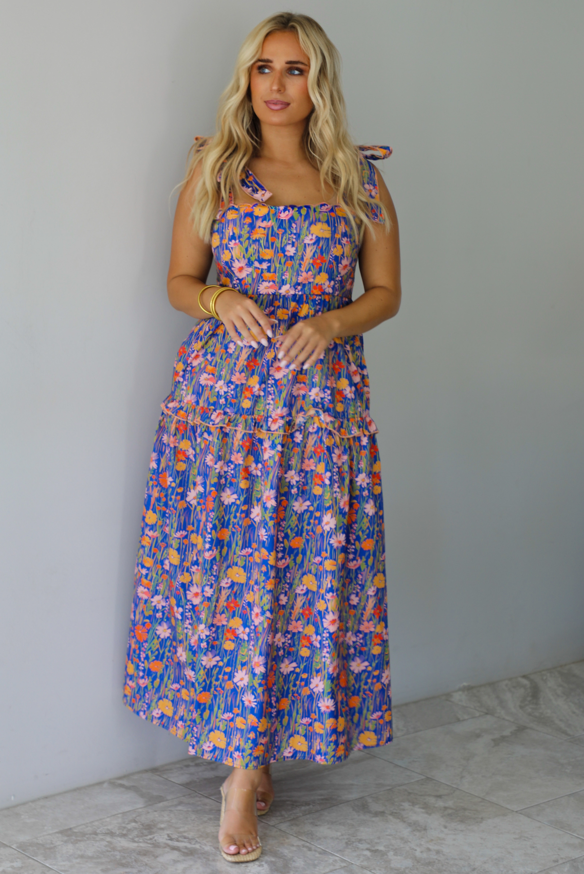A woman wearing a sleeveless tiered navy blue maxi dress with a colorful floral print with a fitted bodice and flowy skirt with functional pockets on either side, standing indoors against a white wall with white marble flooring. 
