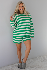 Woman wearing a green and white striped set featuring an oversized long sleeved top and high waisted shorts. The photo is taken indoors against a white wall with white marble flooring. 