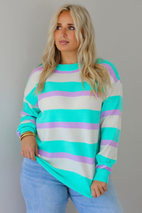 A woman wearing a turquoise, lavender, and cream striped knit sweater, untucked to show the full length, paired with light wash denim jeans, standing indoors against a white wall.
