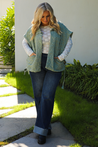 Woman model wearing a sage colored oversized vest paired with a white lace long sleeved top and dark denim outdoors.  The vest has functional pockets on either side, functional button closures in the front, and an oversized fit. 