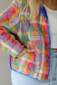 Woman wearing a multi-colored plaid, quilted style jacket paired with a white top underneath and high waisted blue lounge pants. The photo shows a closeup of the jacket, with the functional button closures in the front, functional pockets, and quilted texture. The photo is taken indoors against a white wall.