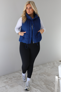 Woman wearing a navy colored puffer vest, white long-sleeve shirt, black leggings, and gray sneakers with white ankle socks indoors against a white wall with white marble flooring.