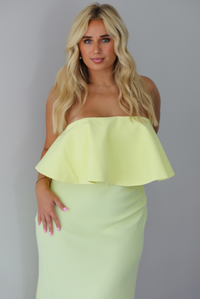 A woman modeling a pale yellow strapless midi dress with a ruffled detail at the top that covers the bodice and a fitted skirt, standing indoors against a white wall. The photo shows a closeup of the bodice. 