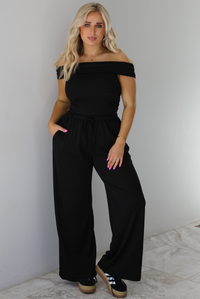 Woman wearing a two piece set featuring an off the shoulder top and high waisted pants. The photo is taken indoors against a white wall with white marble flooring. 