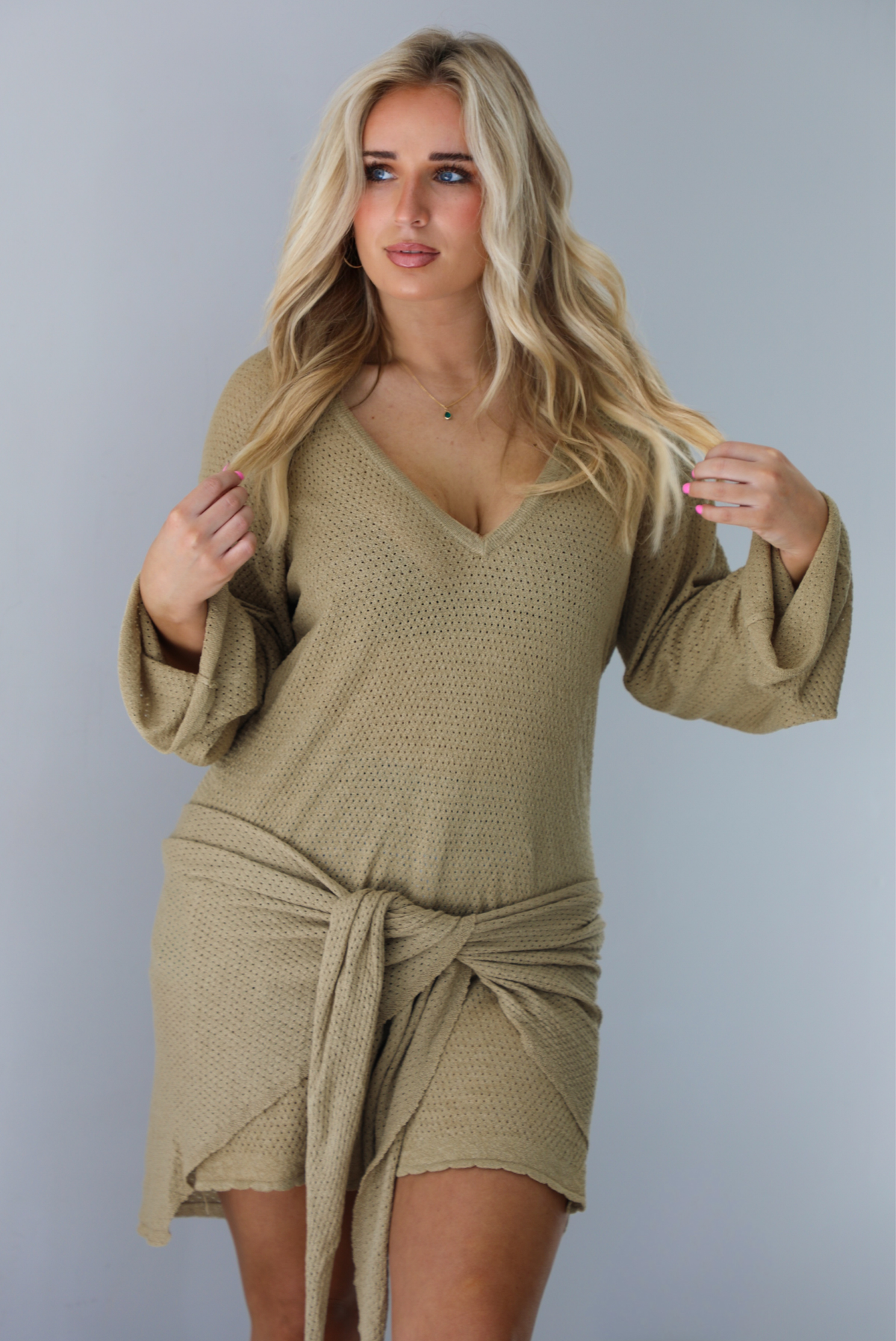 Woman wearing a camel long-sleeve dress that has a tie detail in the front.  The photo is taken indoors against a white wall.