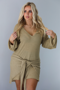Woman wearing a camel long-sleeve dress that has a tie detail in the front.  The photo is taken indoors against a white wall.