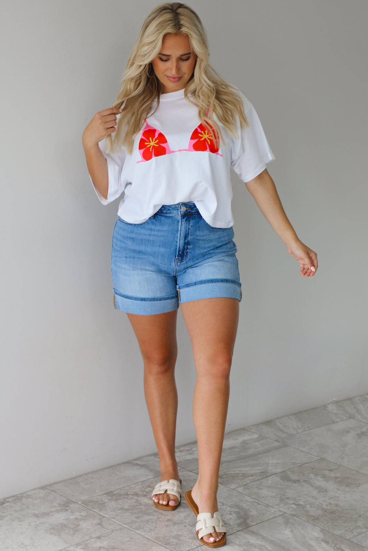 Woman wearing a white cropped boxy tee with a pink, red, and yellow bikini graphic on the front with a pair of medium wash denim shorts indoors against a white wall with white marble flooring.