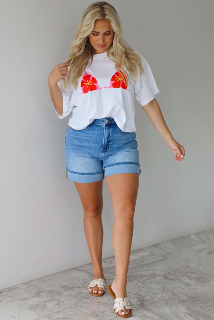 Woman wearing a white cropped boxy tee with a pink, red, and yellow bikini graphic on the front with a pair of medium wash denim shorts indoors against a white wall with white marble flooring.