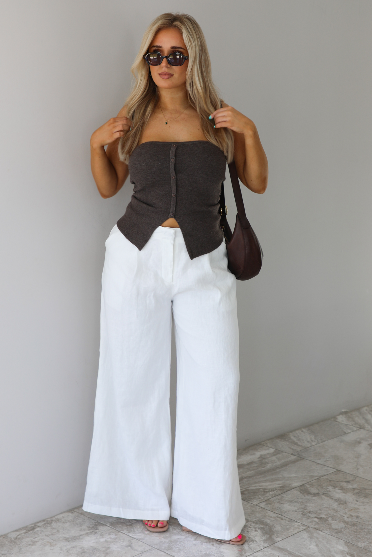 Woman wearing a strapless brown top with non-functional button closures in the front with  a split hem detail, and high waisted, flared hem white linen pants with a brown shoulder bag. The photo is taken indoors against a white wall with white marble flooring. 