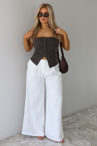 Woman wearing a strapless brown top with non-functional button closures in the front with  a split hem detail, and high waisted, flared hem white linen pants with a brown shoulder bag. The photo is taken indoors against a white wall with white marble flooring. 