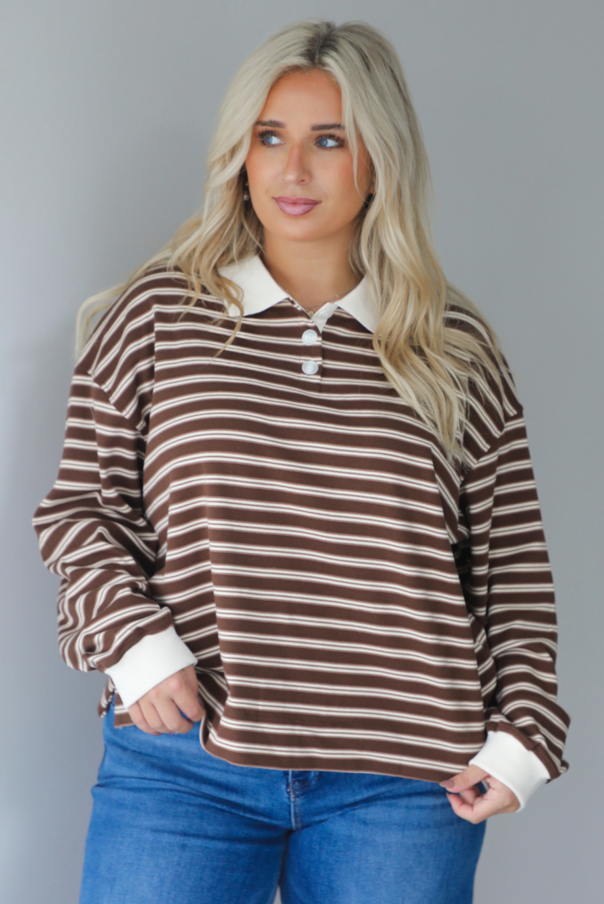 A woman wearing a brown and cream striped collared quarter-button top, paired with dark, wide leg denim with a cuffed hem, standing indoors against a white wall. The top is untucked to show the full length. 