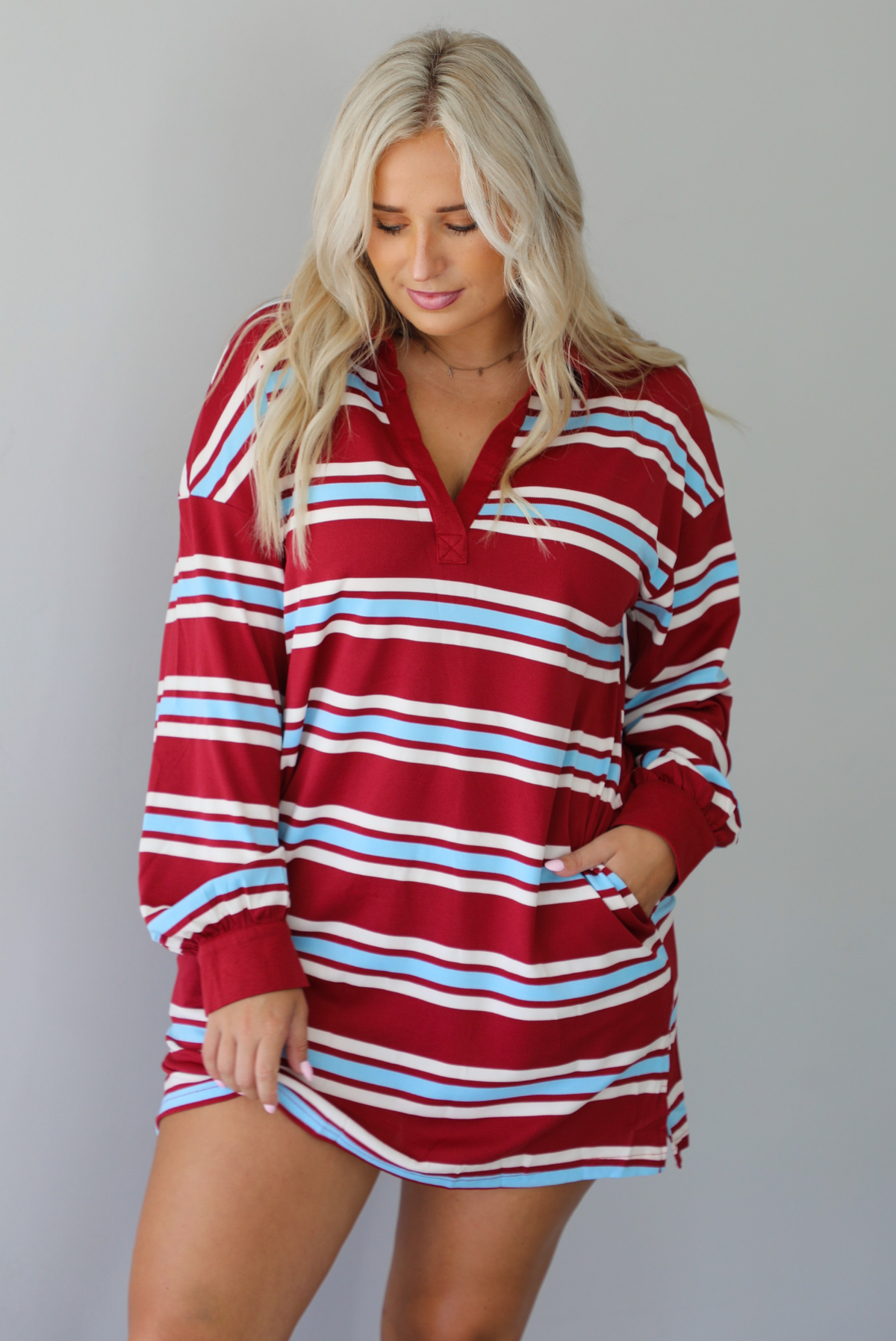 A woman wearing a red burgundy mini dress with a white and blue striped pattern throughout, collared neckline, long sleeves, and functional pockets on ether side, standing indoors against a white wall.