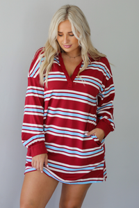 A woman wearing a red burgundy mini dress with a white and blue striped pattern throughout, collared neckline, long sleeves, and functional pockets on ether side, standing indoors against a white wall.