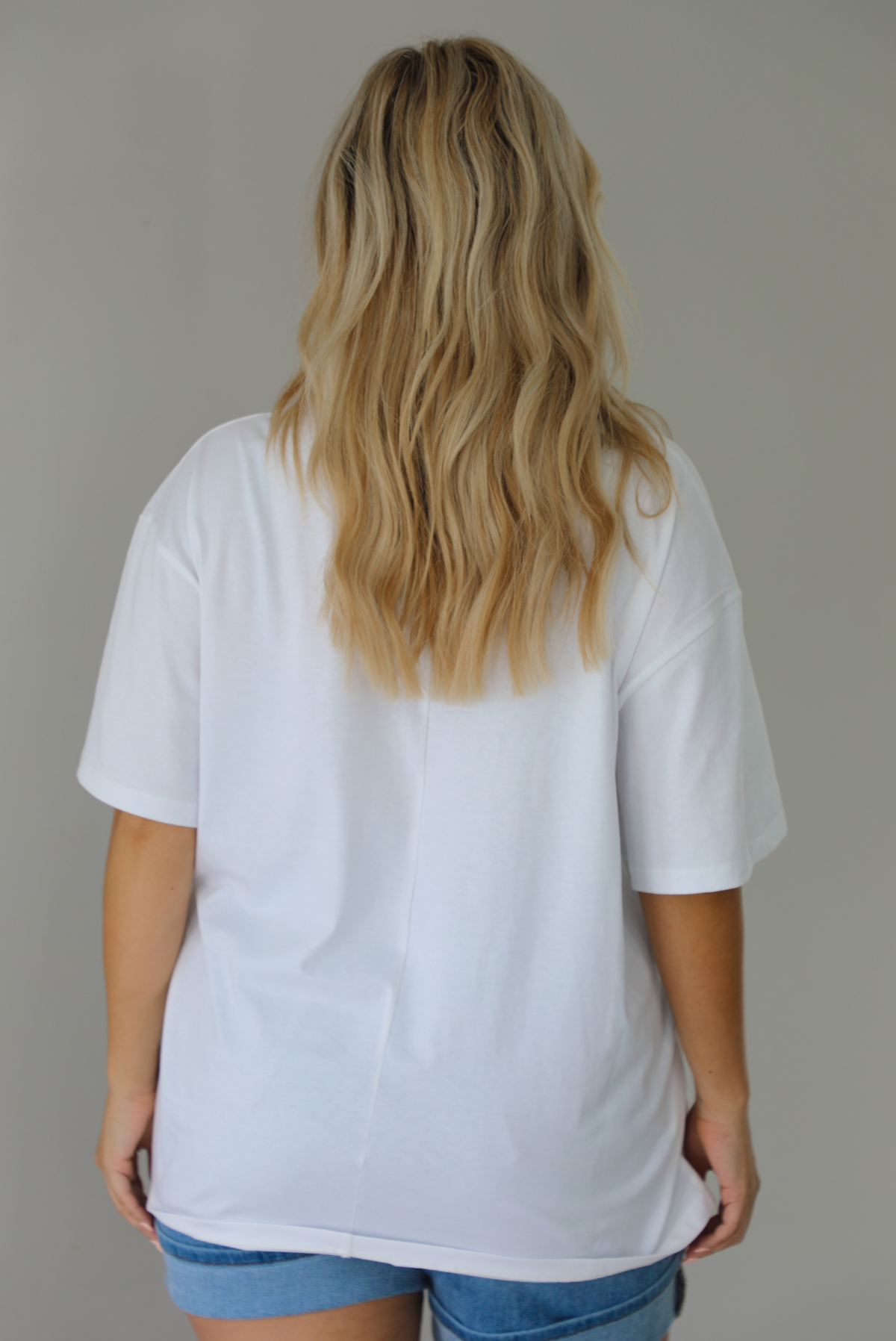Woman wearing a white oversized tee untucked with medium wash denim shorts indoors against a white wall and shows the back of the tee. 