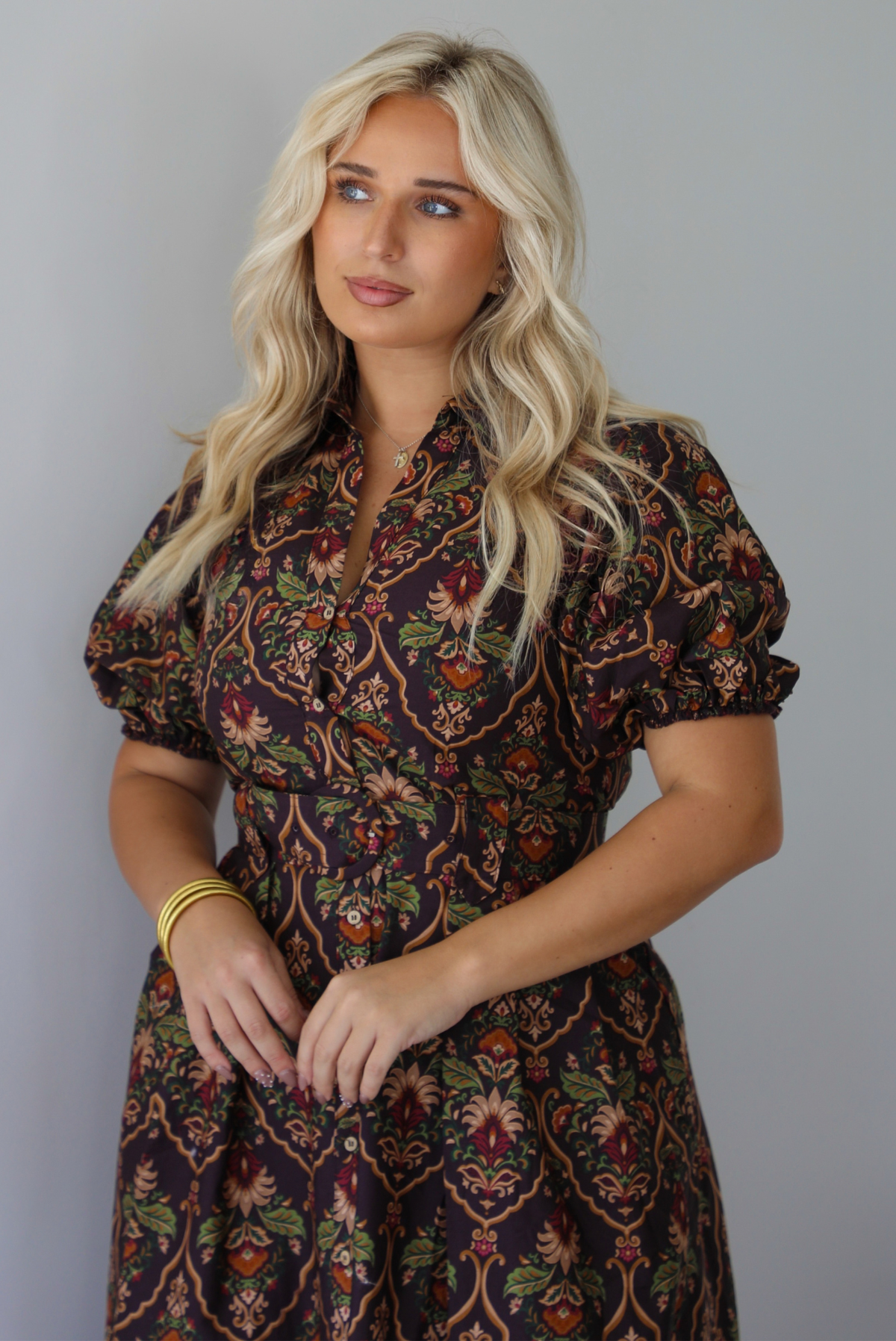 A woman wearing a dark brown midi dress with a multi-colored design throughout, a collared neckline, short puffed sleeves, functional button closures along the front, a belted detail at the waistline, standing indoors against a white wall. The photo shows a closeup of the bodice of the dress. 