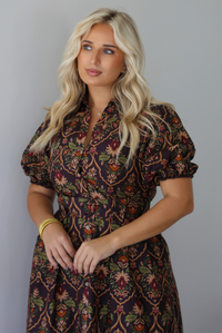 A woman wearing a dark brown midi dress with a multi-colored design throughout, a collared neckline, short puffed sleeves, functional button closures along the front, a belted detail at the waistline, standing indoors against a white wall. The photo shows a closeup of the bodice of the dress. 