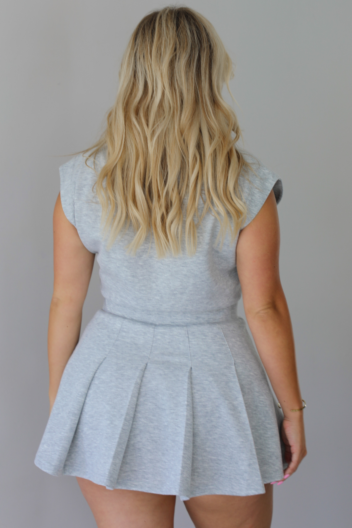 A woman modeling a light gray heathered short-sleeve collared dress with a pleated mini skirt,  indoors against a white wall. The photo shows the back of the dress. 
