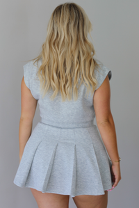 A woman modeling a light gray heathered short-sleeve collared dress with a pleated mini skirt,  indoors against a white wall. The photo shows the back of the dress. 