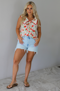Woman wearing a cream and red strawberry patterned top with a collared detail and functional button closures in the front with light wash denim shorts indoors against a white wall with white marble flooring. 