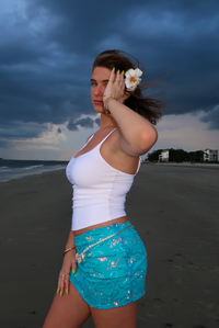 A woman stands on a beach wearing a white ribbed tank top and a bright turquoise sequined wrap skirt with a white floral hair accessory, set against a dark stormy sky.