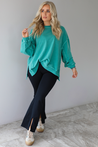Woman wearing a teal oversized sweater with black leggings that have a slightly flared hem with split hem details in the front. The photo is taken in front of a white wall with white marble flooring. 