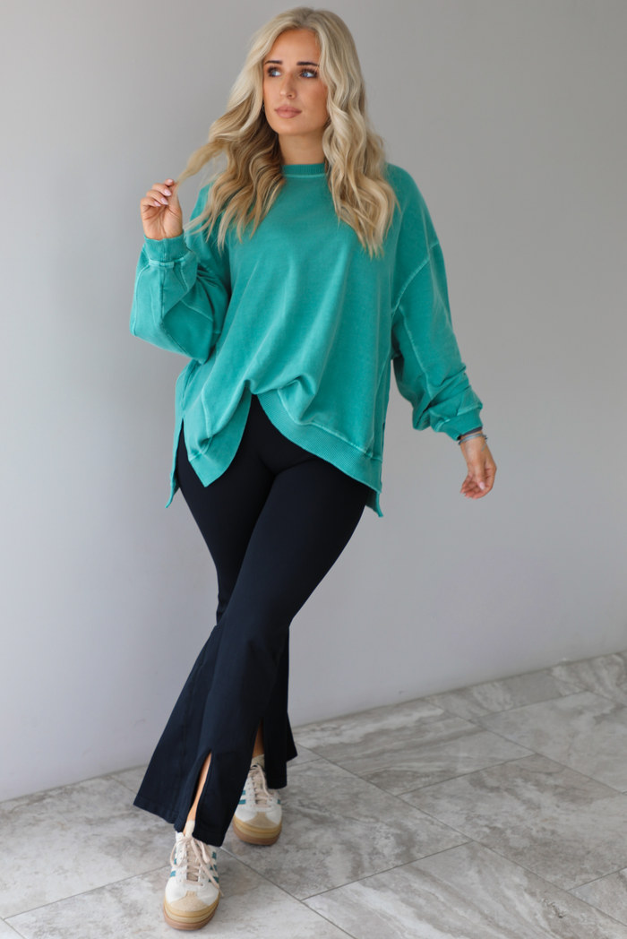 Woman wearing a teal oversized sweater with black leggings that have a slightly flared hem with split hem details in the front. The photo is taken in front of a white wall with white marble flooring. 
