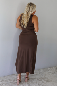 A woman with blonde hair is shown from behind wearing a sleeveless, dark brown set and nude block-heel sandals, standing indoors against a white wall with white marble flooring. 