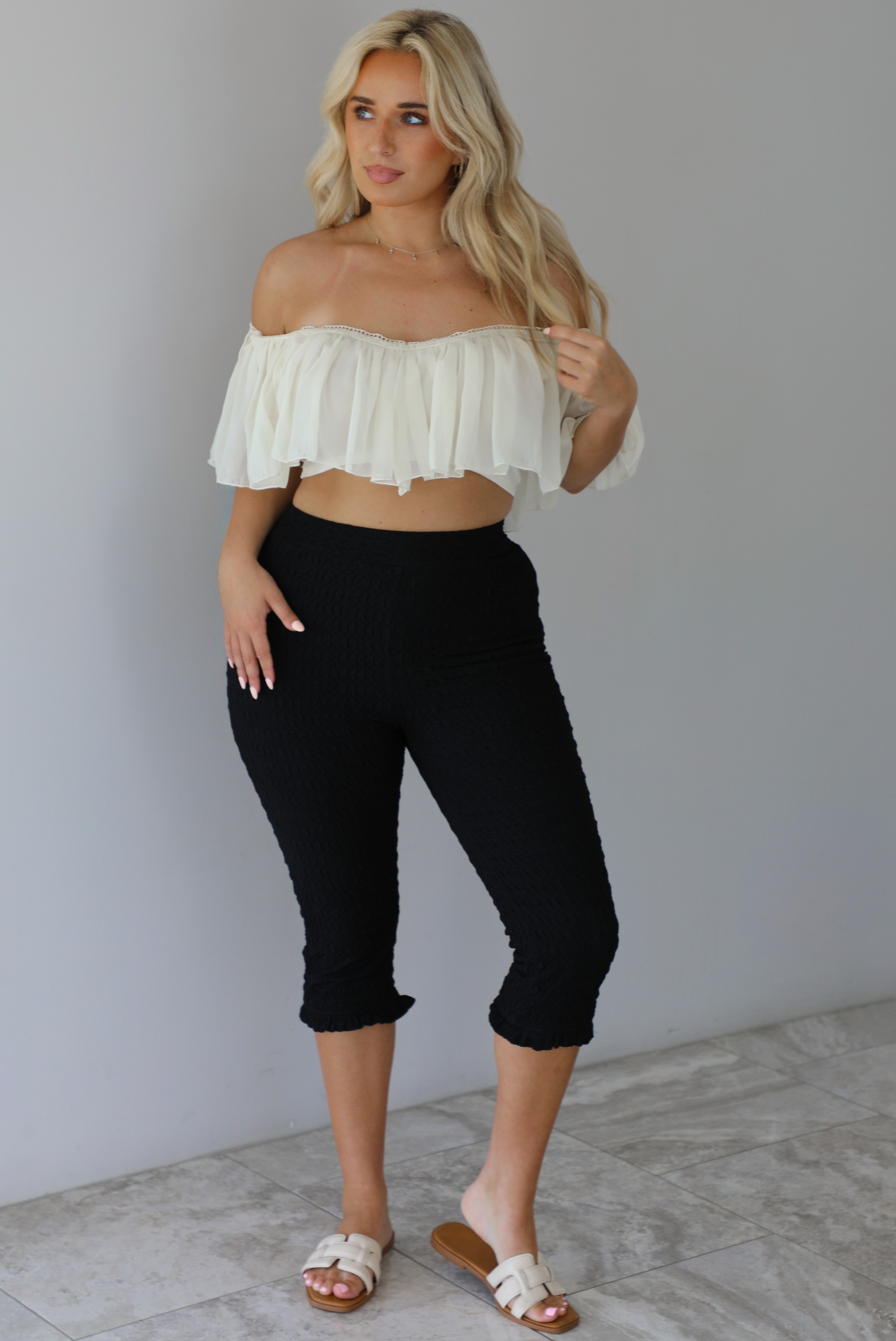 Woman wearing a cream ruffled cropped top with high waisted black, textured capri pants indoors against a white wall with white marble flooring. The photo shows the top being worn off the shoulder. 