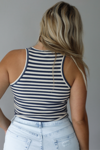 Woman wearing a navy and white striped cropped tank top with beaded crab graphic on the front with light wash denim shorts indoors against a white wall. The photo shows the back of the tank. 