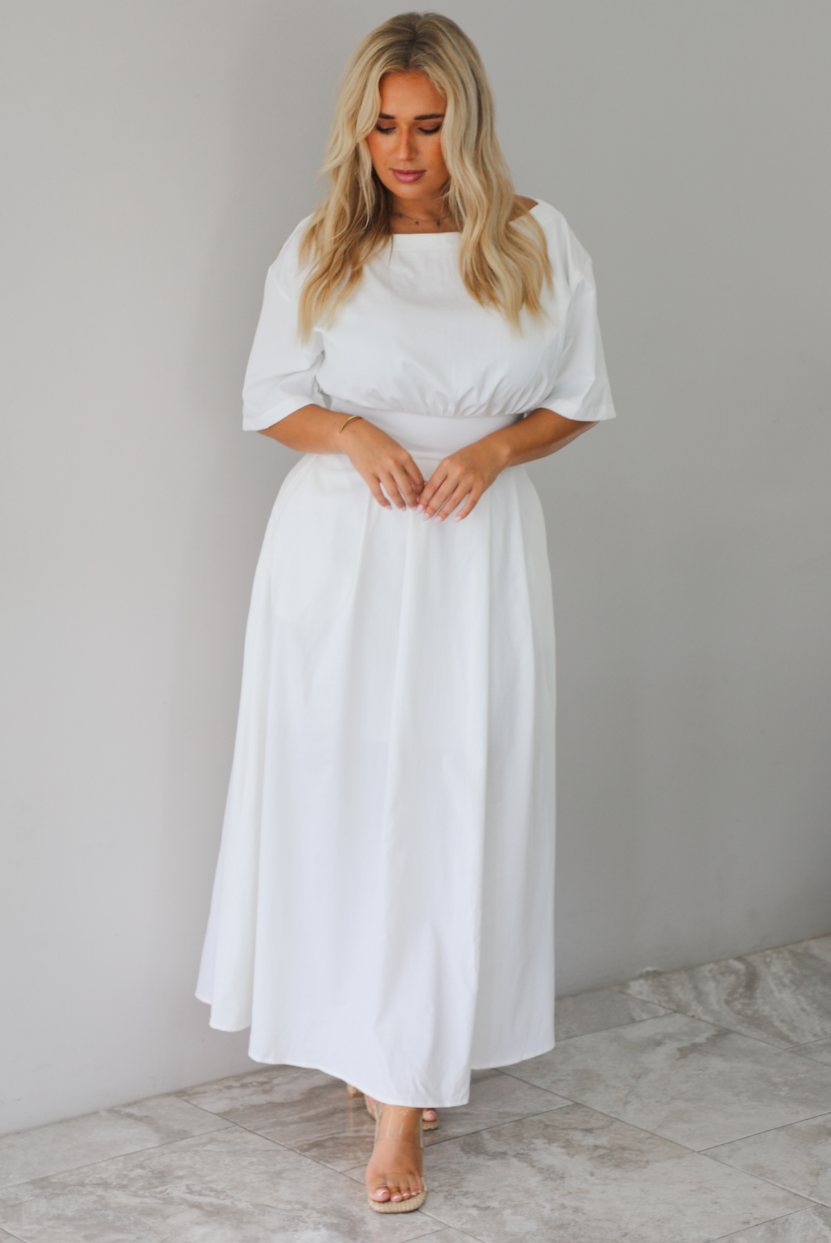 A woman modeling a white midi dress with short puff sleeves, a wide boat neckline, and side pockets, standing indoors against a white wall with white marble flooring. 