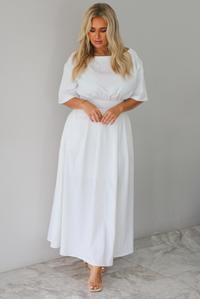 A woman modeling a white midi dress with short puff sleeves, a wide boat neckline, and side pockets, standing indoors against a white wall with white marble flooring. 