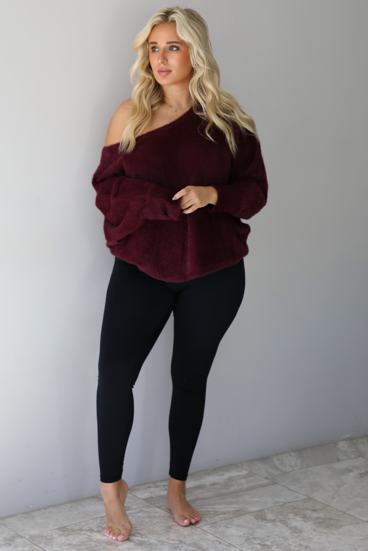 A woman wearing a maroon oversized long sleeved sweater, worn off the shoulder, paired with black leggings, indoors against a white wall with white marble flooring.